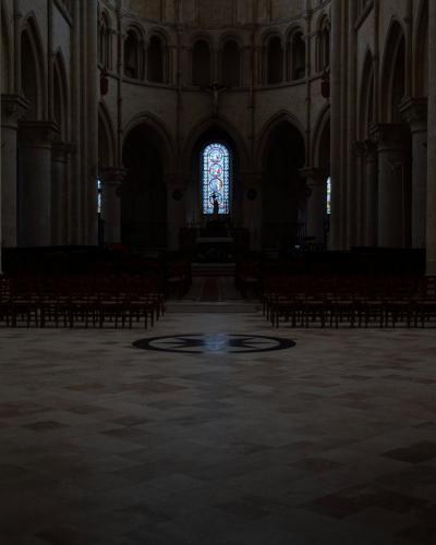 1. The image shows the dimly lit interior nave of a large stone church, featuring tall columns, arched windows, and rows of wooden chairs facing an altar area with a stained-glass window at the far end.