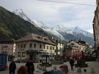 1. A town square in a mountainous alpine setting featuring multi-story buildings, a central fountain, pedestrians, and snow-capped peaks in the background.