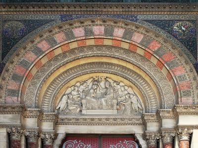 A close-up architectural detail of an ornate stone archway featuring a sculpted tympanum with religious figures, surrounded by decorative mosaics and striped stonework.
