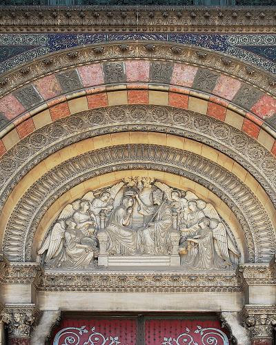 A close-up architectural detail of an ornate stone archway featuring a sculpted tympanum with religious figures, surrounded by decorative mosaics and striped stonework.