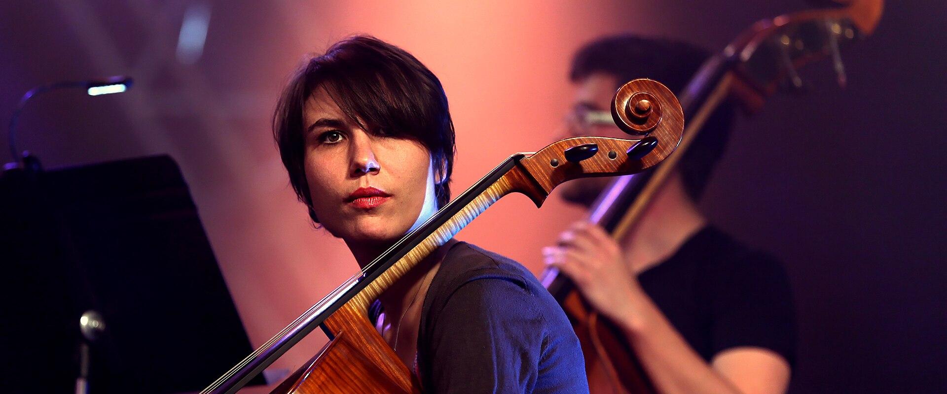 A female musician with short dark hair plays a cello on a dimly lit stage under purple and orange spotlights, with another musician playing a double bass in the background.