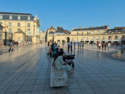 The image shows a large, paved public square with classical European architecture, including a prominent building with a mansard roof on the left and a curved arcade building on the right, with people walking and sitting on benches under a clear b