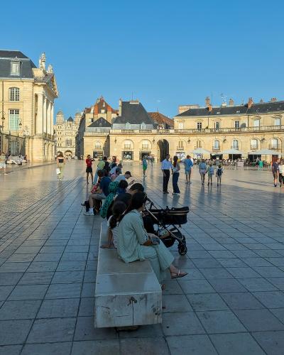 The image shows a large, paved public square with classical European architecture, including a prominent building with a mansard roof on the left and a curved arcade building on the right, with people walking and sitting on benches under a clear b