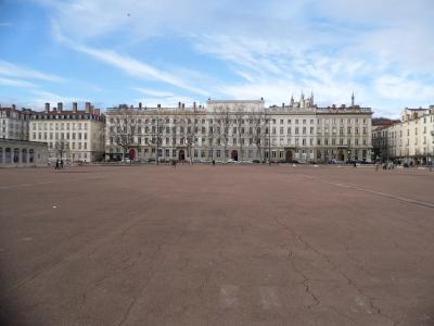 A wide public square with a distinctive red ground surface, featuring a large bronze equestrian statue on a stone pedestal in the center, with multi-story buildings and a hillside in the background.