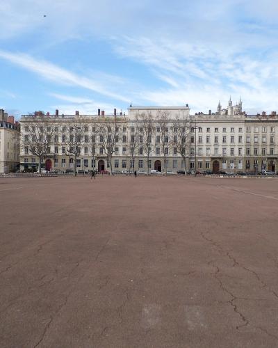 A wide public square with a distinctive red ground surface, featuring a large bronze equestrian statue on a stone pedestal in the center, with multi-story buildings and a hillside in the background.
