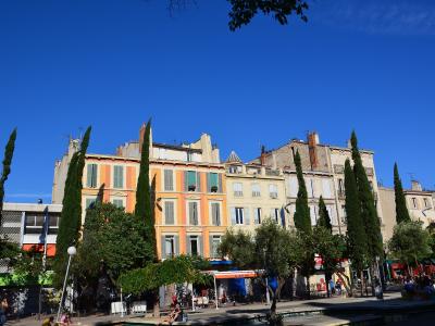 A sunlit urban square lined with colorful multi-story buildings featuring shuttered windows, interspersed with tall cypress trees and leafy green trees; people are seated at outdoor cafes and walking along the paved plaza under a clear blue sky.