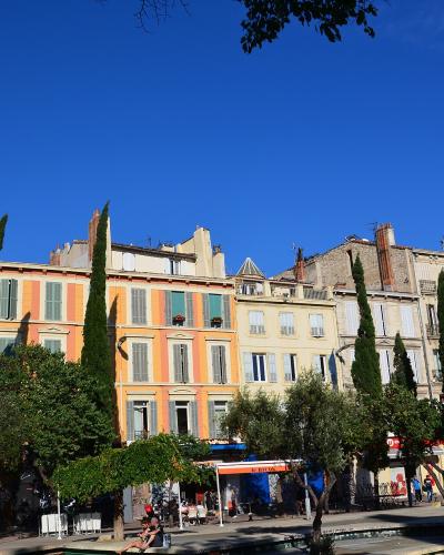 A sunlit urban square lined with colorful multi-story buildings featuring shuttered windows, interspersed with tall cypress trees and leafy green trees; people are seated at outdoor cafes and walking along the paved plaza under a clear blue sky.