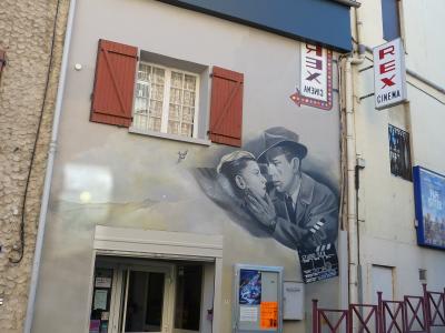 The image shows the exterior of a cinema building with a large mural of a man and woman, signage reading "CINEMA REX" and "STUDIO REX," and a vertical sign also reading "REX CINEMA."