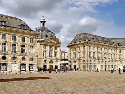 A wide-angle view of a grand, symmetrical 18th-century European square with classical stone buildings, arched ground floors, and a central pavilion topped by a dome and spire; people are walking or sitting on benches across the cobblestone plaza u