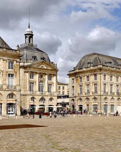 A wide-angle view of a grand, symmetrical 18th-century European square with classical stone buildings, arched ground floors, and a central pavilion topped by a dome and spire; people are walking or sitting on benches across the cobblestone plaza u