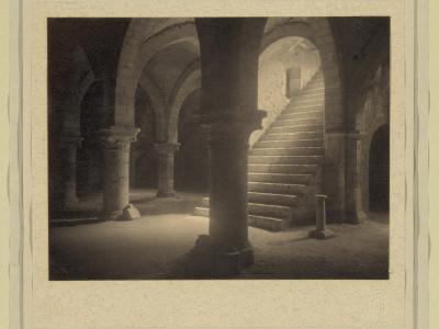 A vintage black-and-white photograph showing a stone crypt or cellar with arched ceilings, thick columns, and a staircase leading upward, mounted on a textured mat with a faint stamp in the lower right corner.