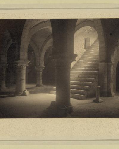 A vintage black-and-white photograph showing a stone crypt or cellar with arched ceilings, thick columns, and a staircase leading upward, mounted on a textured mat with a faint stamp in the lower right corner.