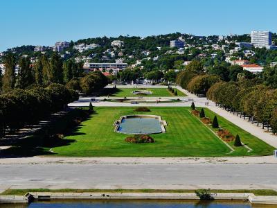 A wide-angle view of a large, formal public park with manicured lawns, symmetrical tree-lined avenues, rectangular ponds, and a hillside cityscape in the background under a clear blue sky.