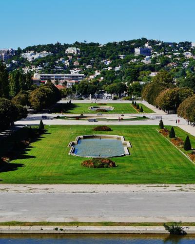 A wide-angle view of a large, formal public park with manicured lawns, symmetrical tree-lined avenues, rectangular ponds, and a hillside cityscape in the background under a clear blue sky.