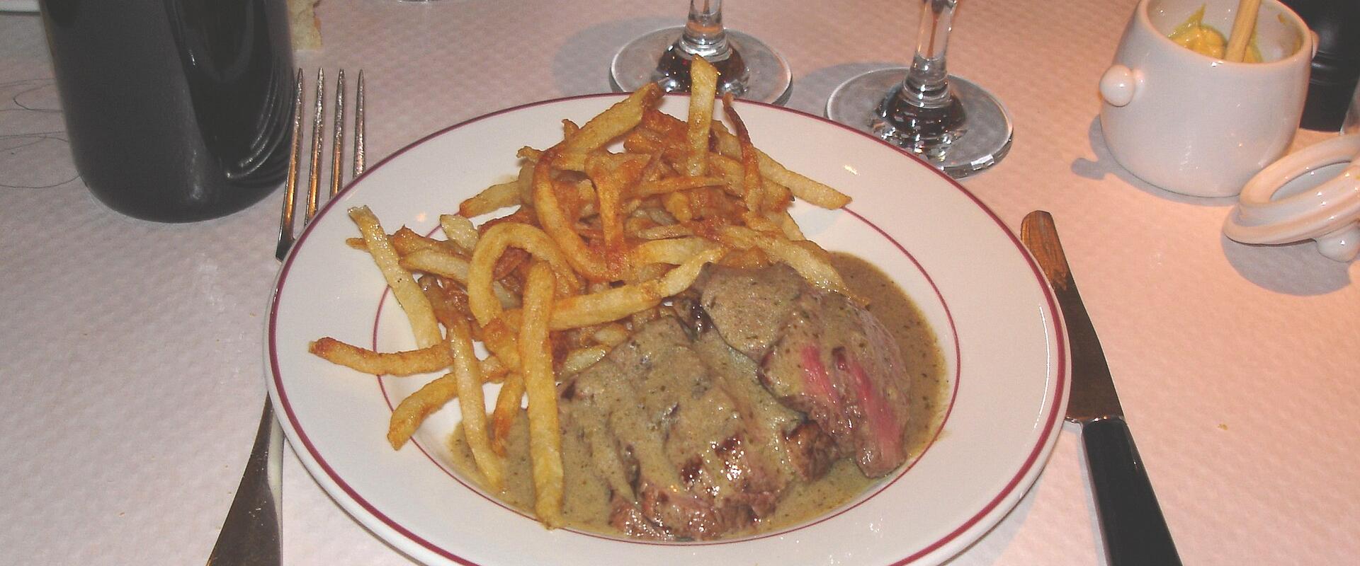 A plate of steak frites with a creamy sauce, served on a white plate with a red rim, accompanied by wine glasses, cutlery, and a small ceramic container with a wooden spoon, likely mustard, on a white tablecloth.