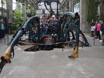 The image shows a large, mechanical spider sculpture with glowing red eyes and riders seated on its back, situated indoors within a structure featuring concrete pillars, glass roofing, and hanging greenery, surrounded by spectators.