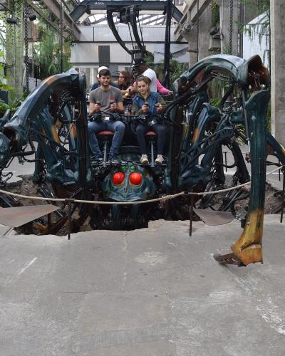 The image shows a large, mechanical spider sculpture with glowing red eyes and riders seated on its back, situated indoors within a structure featuring concrete pillars, glass roofing, and hanging greenery, surrounded by spectators.
