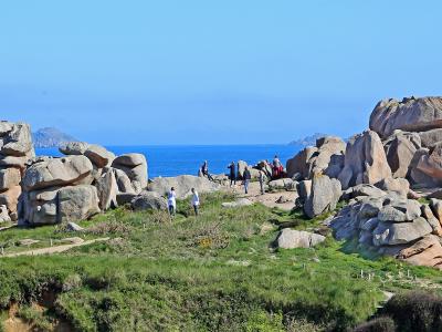 A coastal landscape featuring large, rounded granite rock formations on a grassy cliff edge overlooking the blue sea, with tourists walking along a path.