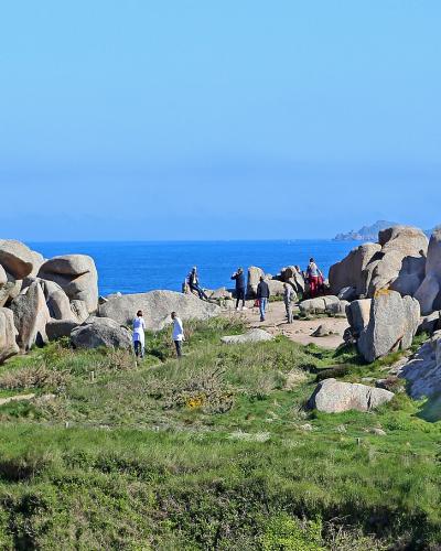 A coastal landscape featuring large, rounded granite rock formations on a grassy cliff edge overlooking the blue sea, with tourists walking along a path.