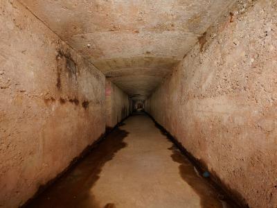 A long, narrow underground tunnel with rough, earthen walls and a damp floor, featuring concrete ceiling segments and faint graffiti or markings on the left wall.