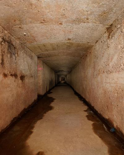 A long, narrow underground tunnel with rough, earthen walls and a damp floor, featuring concrete ceiling segments and faint graffiti or markings on the left wall.