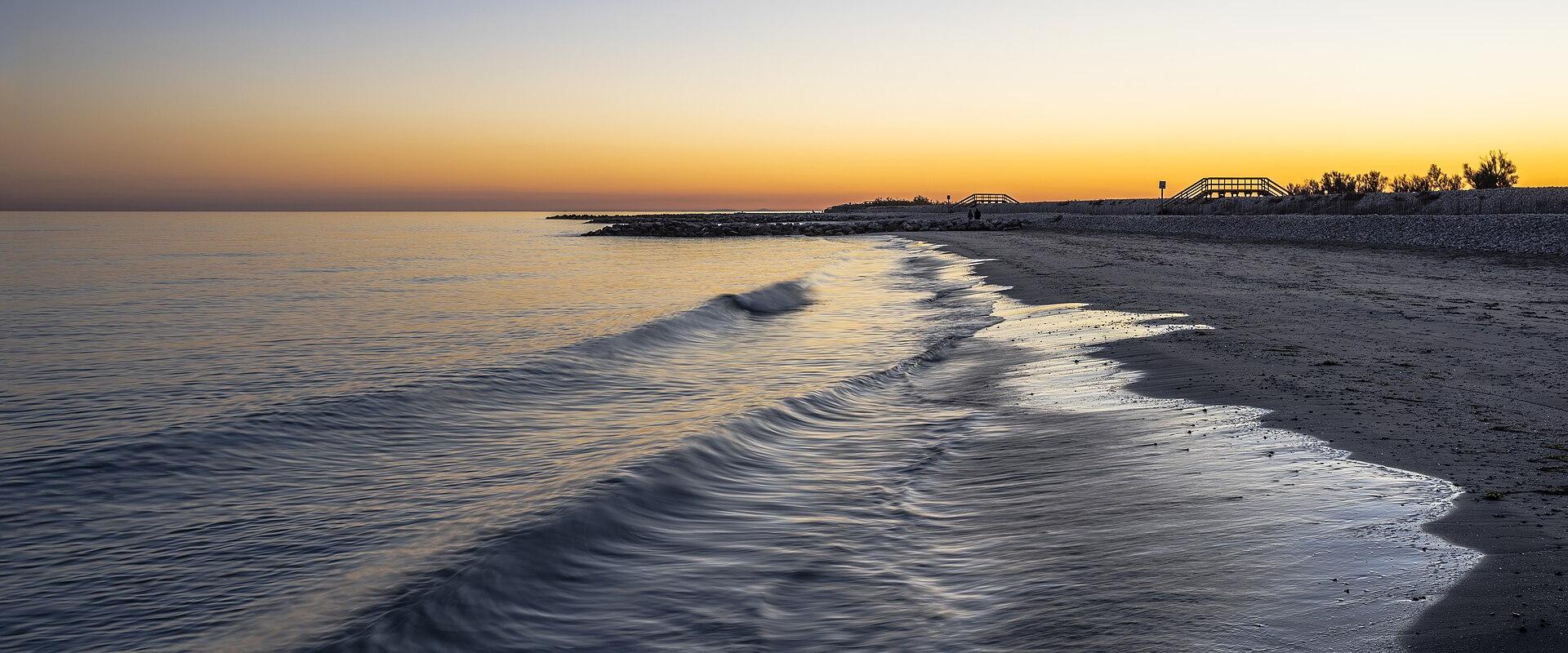 A long-exposure photograph of a calm beach at sunset, showing gentle waves rolling onto wet sand, with a stone breakwater and a metal pedestrian bridge visible in the distance along the shoreline.