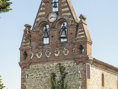 A brick and stone bell tower with a triangular gable, three bells, a clock face, and a cross on top, surrounded by green foliage under a clear sky.
