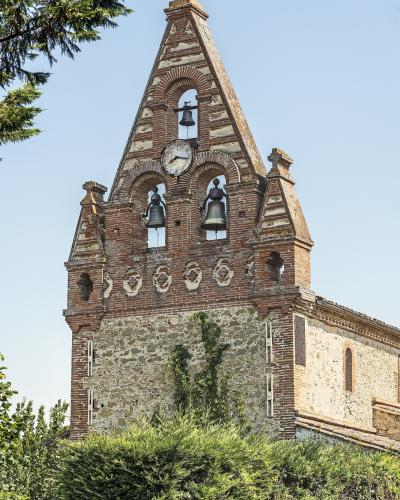 A brick and stone bell tower with a triangular gable, three bells, a clock face, and a cross on top, surrounded by green foliage under a clear sky.