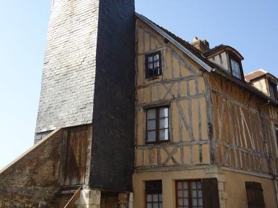 A tall, narrow clock tower clad in dark grey slate tiles stands adjacent to a half-timbered house with yellow stucco and wooden beams, under a clear blue sky.