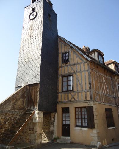 A tall, narrow clock tower clad in dark grey slate tiles stands adjacent to a half-timbered house with yellow stucco and wooden beams, under a clear blue sky.