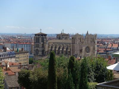 A wide-angle view of a large medieval cathedral with two towers, flying buttresses, and rose windows, set against a dense urban backdrop of red-tiled roofs and distant hills under a clear sky.
