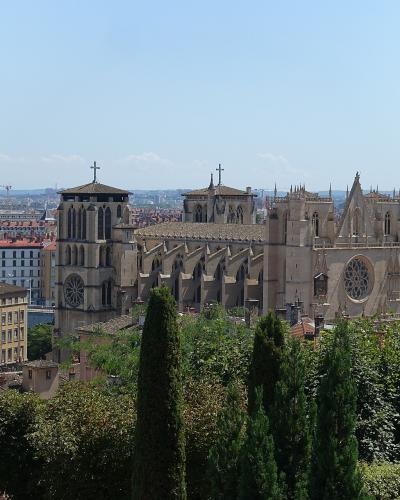 A wide-angle view of a large medieval cathedral with two towers, flying buttresses, and rose windows, set against a dense urban backdrop of red-tiled roofs and distant hills under a clear sky.