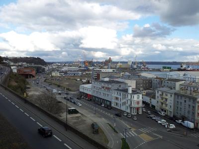A high-angle view of an urban waterfront area in France, featuring a road with traffic, a large white building with "CAP OUEST" signage, and a background of industrial port facilities with cranes and ships.