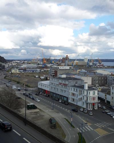 A high-angle view of an urban waterfront area in France, featuring a road with traffic, a large white building with "CAP OUEST" signage, and a background of industrial port facilities with cranes and ships.