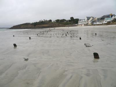 The image shows a wide, wet sandy beach at low tide with rows of dark, blocky concrete anti-tank obstacles (Czech hedgehogs or similar Wehrmacht-era defenses) protruding from the sand, with houses and a grassy headland in the background under an o