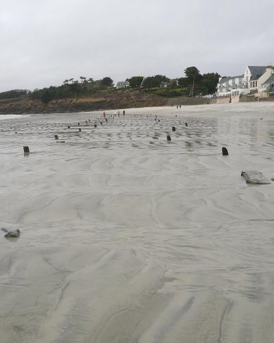 The image shows a wide, wet sandy beach at low tide with rows of dark, blocky concrete anti-tank obstacles (Czech hedgehogs or similar Wehrmacht-era defenses) protruding from the sand, with houses and a grassy headland in the background under an o