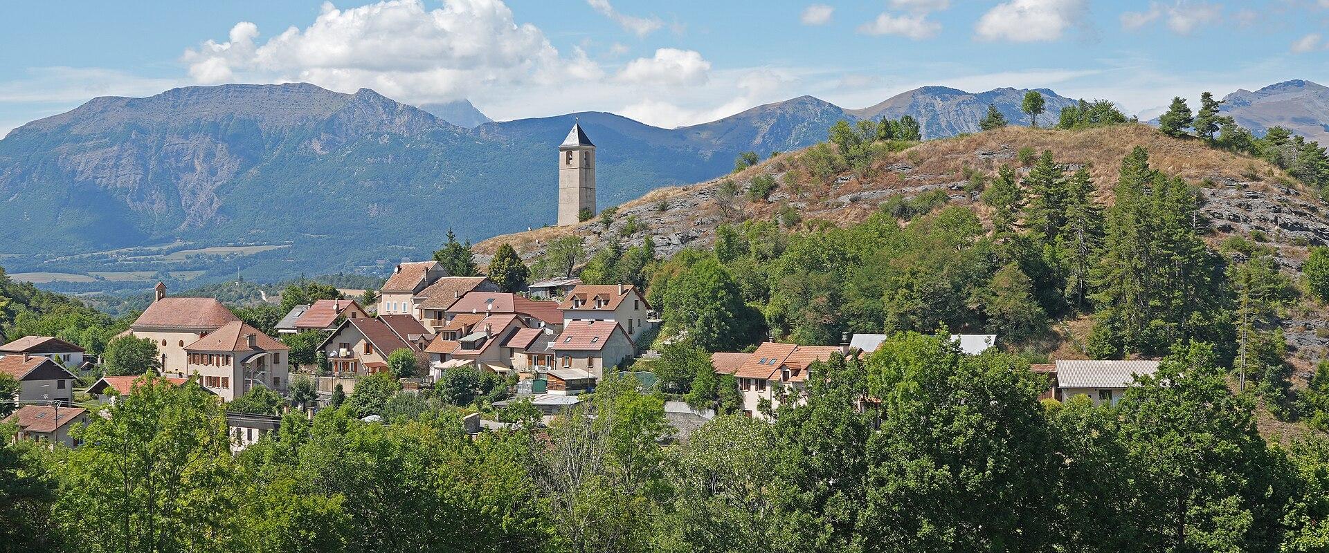 A sunlit hillside village with terracotta-roofed houses clustered around a prominent stone bell tower, set against a backdrop of rugged mountains under a blue sky with scattered clouds; foreground shows green fields and dense trees.