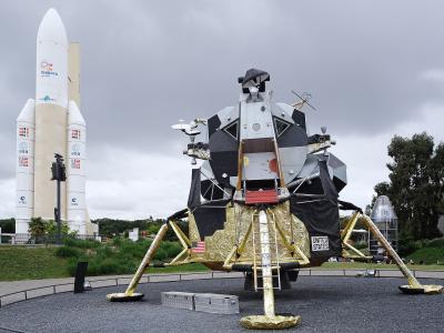 The image shows a full-scale replica of the Apollo Lunar Module in the foreground and a large Ariane 5 rocket model in the background, with the "Cité de l'espace" logo visible on the rocket's side.
