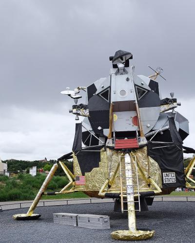The image shows a full-scale replica of the Apollo Lunar Module in the foreground and a large Ariane 5 rocket model in the background, with the "Cité de l'espace" logo visible on the rocket's side.