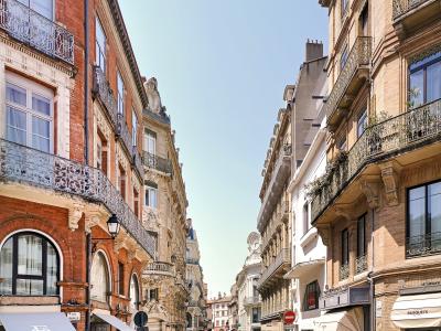 A sunlit, narrow pedestrian street lined with multi-story brick and stone buildings featuring wrought-iron balconies, shop awnings (including one labeled “BUSQUETS”), parked bicycles, and a “No Entry” sign; the street nameplate on the left reads “