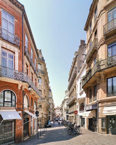 A sunlit, narrow pedestrian street lined with multi-story brick and stone buildings featuring wrought-iron balconies, shop awnings (including one labeled “BUSQUETS”), parked bicycles, and a “No Entry” sign; the street nameplate on the left reads “