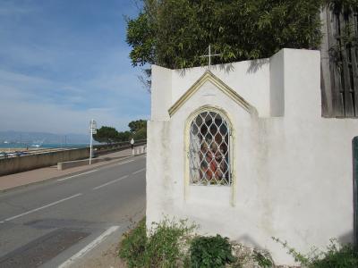 A white roadside religious shrine (or niche) with a cross on top and a statue behind a metal grille, situated next to a paved road with a view of the sea and distant mountains in the background.