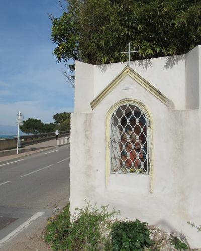A white roadside religious shrine (or niche) with a cross on top and a statue behind a metal grille, situated next to a paved road with a view of the sea and distant mountains in the background.