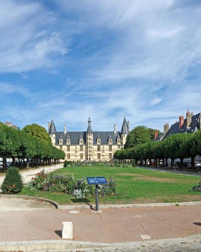 A wide-angle view of a grand Renaissance-style palace with a pale ochre facade, multiple conical-roofed towers, and symmetrical wings, set behind a manicured garden with tree-lined alleys and informational plaques under a blue sky.