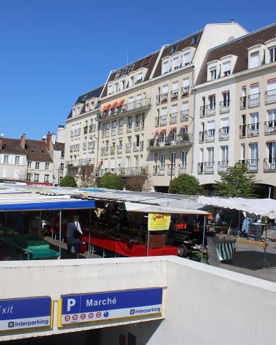 The image shows an outdoor market with stalls under white and blue canopies, situated in a town square surrounded by multi-story European-style buildings, with a parking garage entrance in the foreground displaying signs reading "Sortie / Exit" an