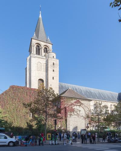 A low-angle photograph of a tall, light-colored stone bell tower with Romanesque architectural features, including arched openings, a steep pyramidal roof topped with a cross, and a clock face on the facade, set against a clear blue sky.