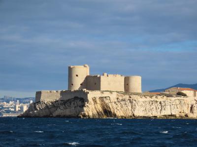 A stone fortress with cylindrical towers and crenellated walls sits atop a rocky island in the sea, with a coastal city skyline visible in the background under a partly cloudy sky.