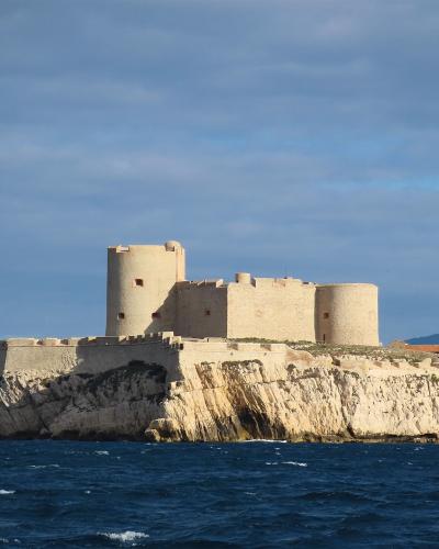 A stone fortress with cylindrical towers and crenellated walls sits atop a rocky island in the sea, with a coastal city skyline visible in the background under a partly cloudy sky.