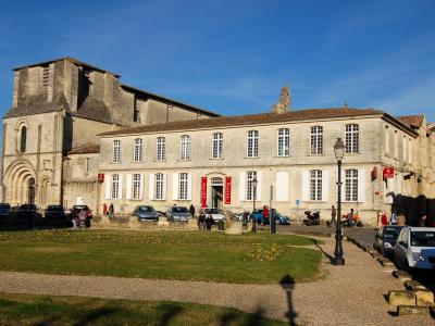 The image shows a large stone building complex in a European town square, featuring a Romanesque church tower on the left and a long, multi-story building with white shutters and red banners to the right, with cars parked in front and people walki