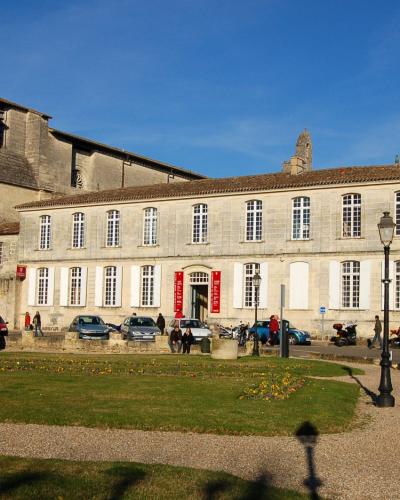 The image shows a large stone building complex in a European town square, featuring a Romanesque church tower on the left and a long, multi-story building with white shutters and red banners to the right, with cars parked in front and people walki